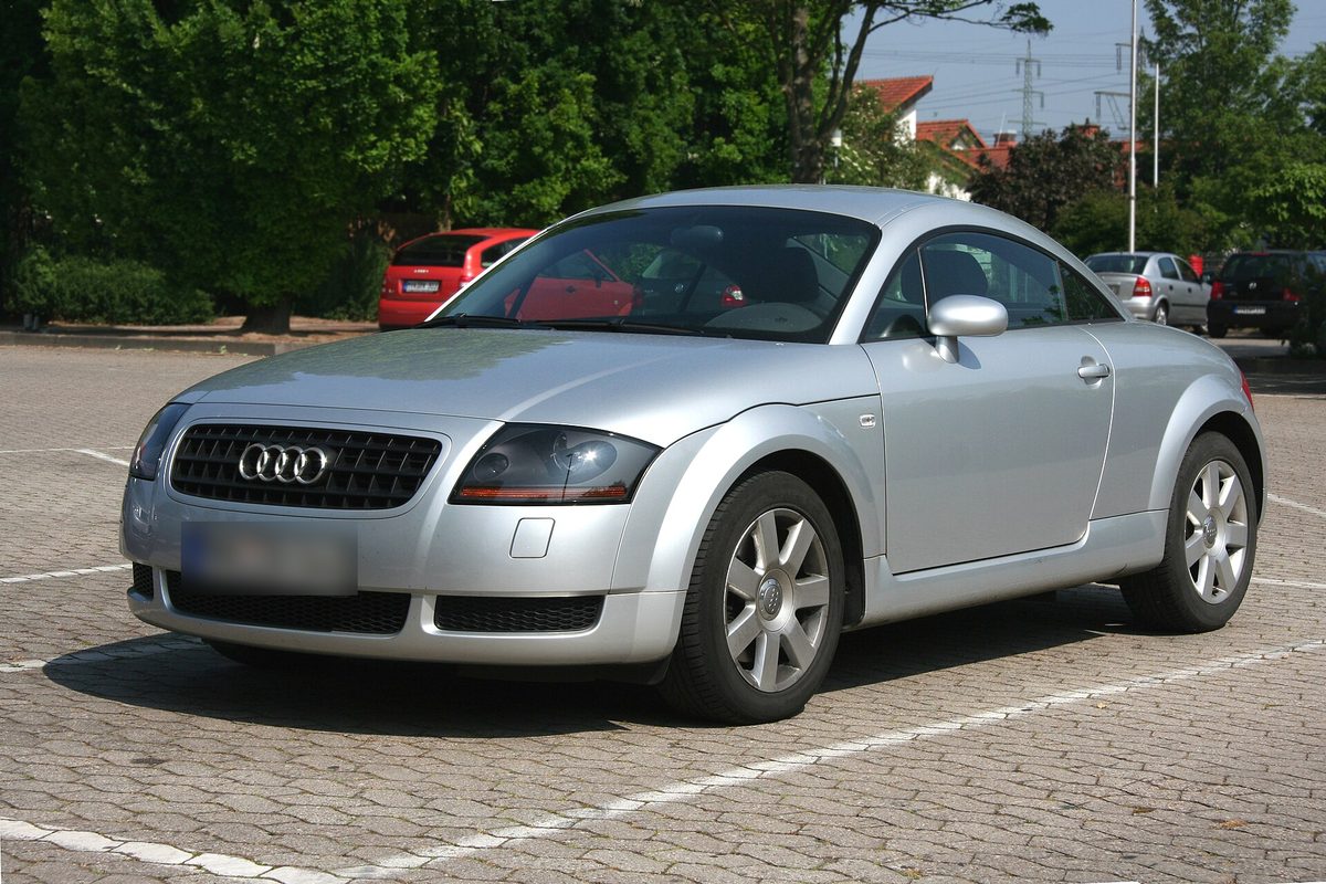Audi TT coupé de primera generación plateado, aparcado en un aparcamiento empedrado con árboles detrás.