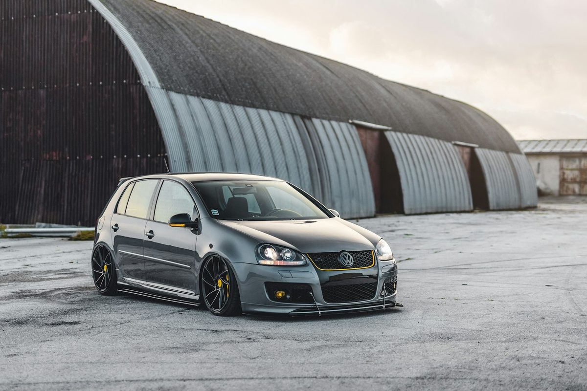Volkswagen Golf GTI de quinta generación gris, rebajado y con llantas no originales, aparcado delante de un hangar.