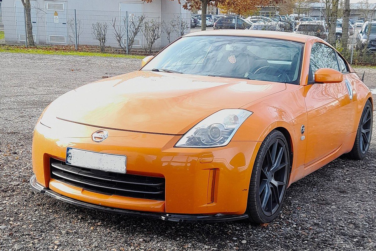 Nissan 350Z coupé naranja con llantas oscuras, aparcado en un aparcamiento de grava.