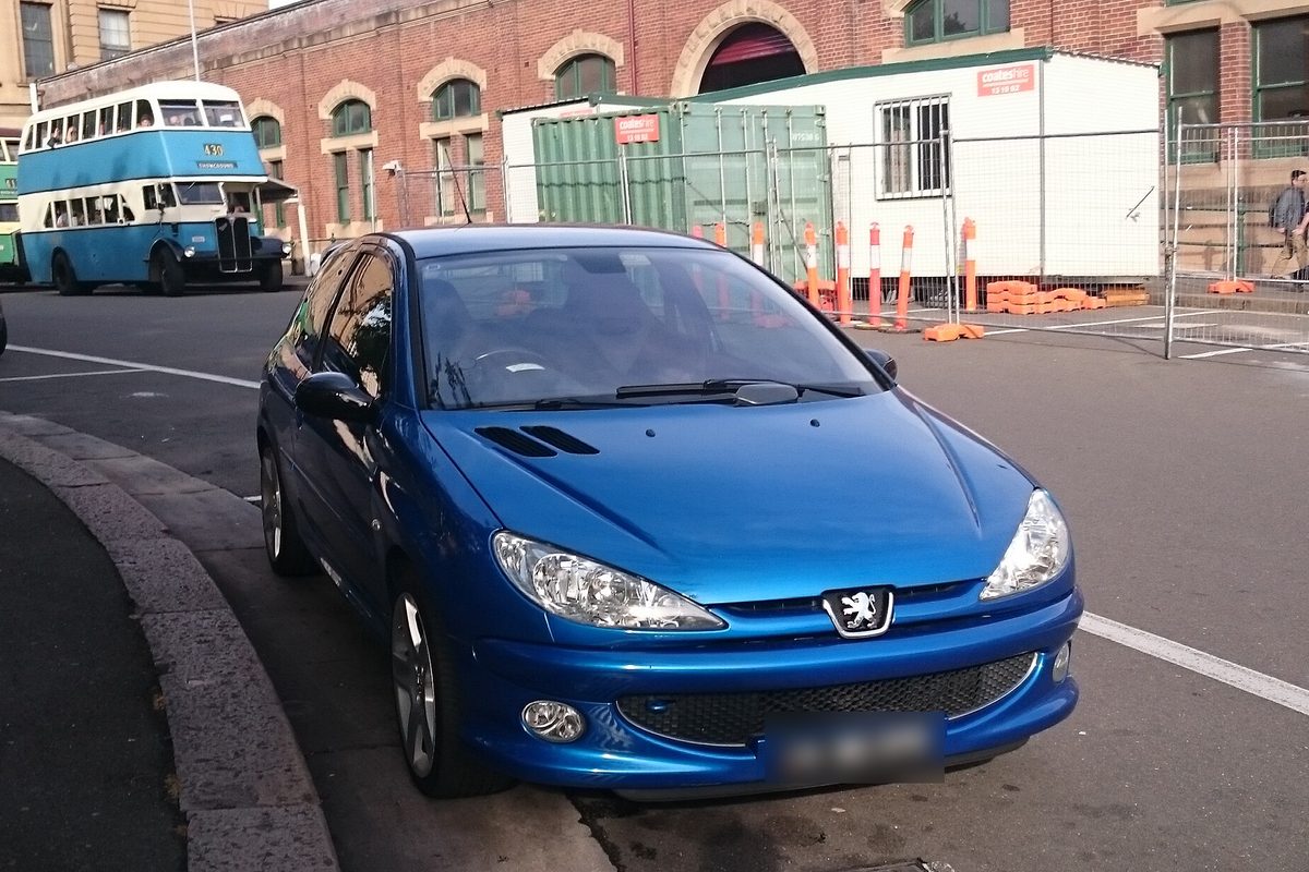 Peugeot 206 GTi 180 azul, de frente, aparcado en una calle con un autobús antiguo detrás.