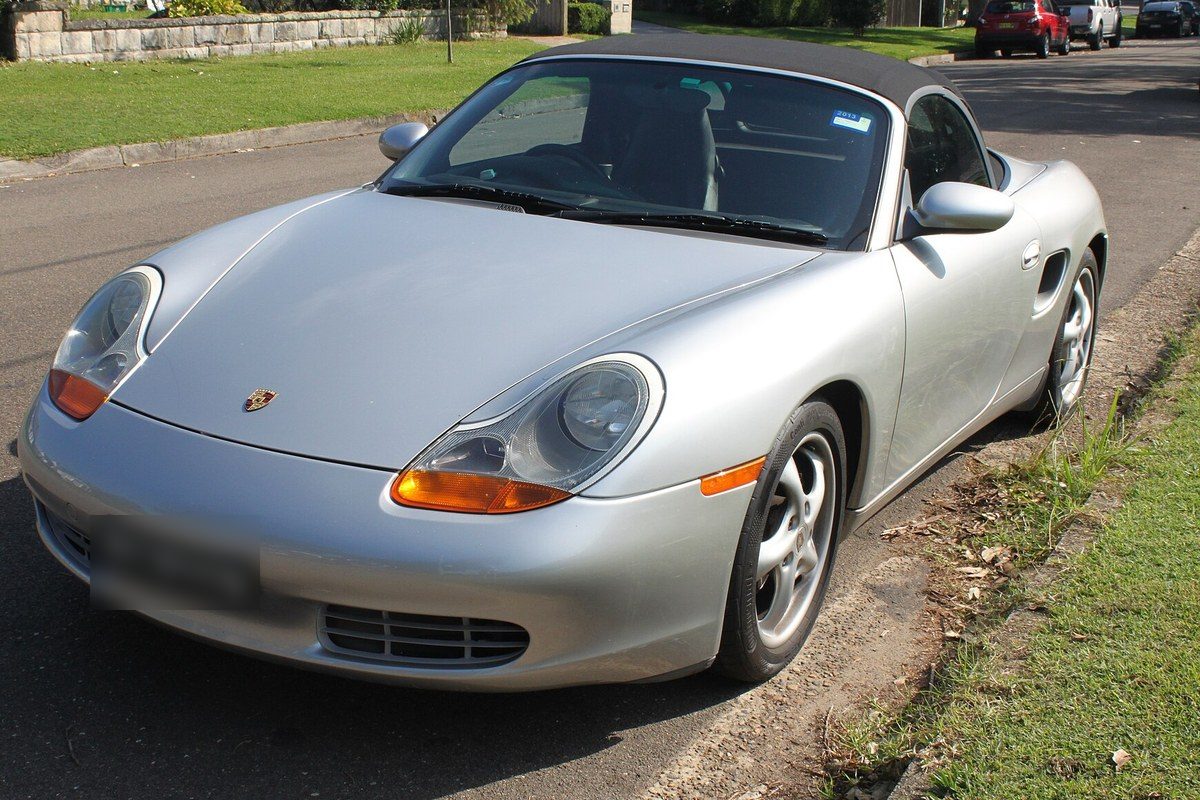 Porsche Boxster de primera generación plateado, con faros anteriores al restyling y la capota negra puesta, aparcado junto a un césped.