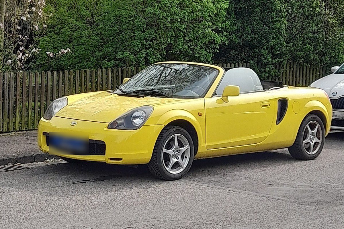 Toyota MR2 Roadster de tercera generación amarillo, descapotado, aparcado en una calle junto a una valla de madera.